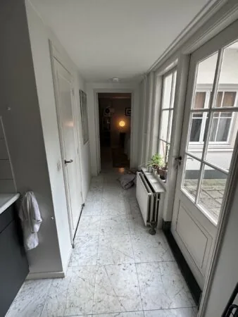 Smalle gang met marmeren vloer, wit geschilderde deuren en een doorkijk naar een verlichte kamer in een woning aan de Eerste Lindendwarsstraat.